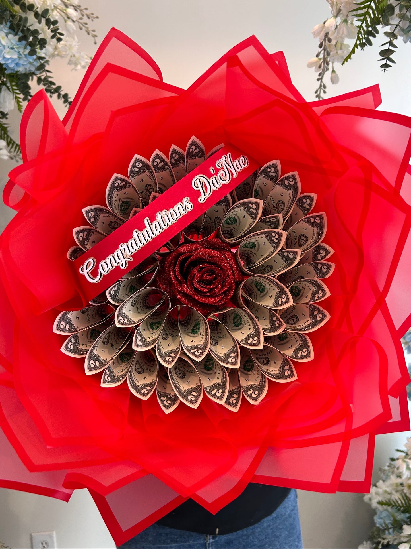A dollar bill flower bouquet with a red rose center, surrounded by folded US dollar bills and wrapped in red sheer paper, with a red ribbon that reads 'Congratulations Beautiful.'.